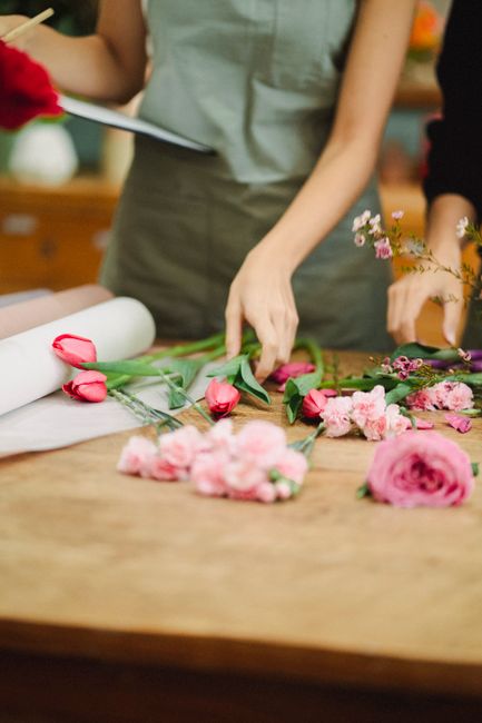 A florist arranging a floral gift at a wooden table, featuring delicate pink roses with lush green stems and leaves, alongside soft pink carnations and small pink and purple blossoms. The flowers are 