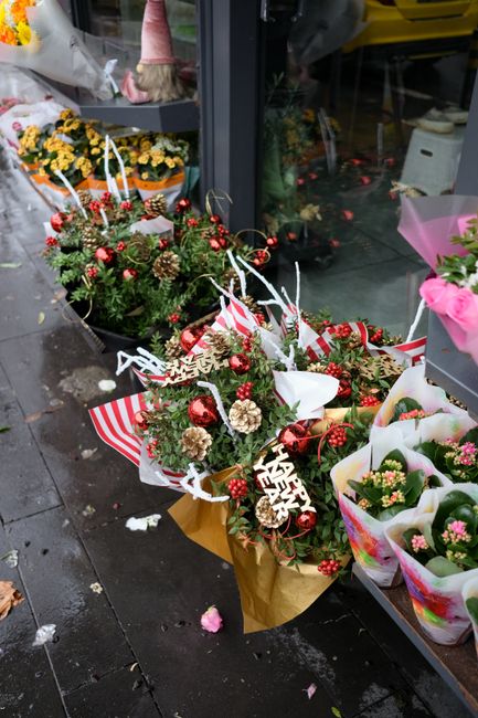 A festive floral arrangement featuring a variety of russet, red, and cream-colored flowers, complemented by green pine branches, red berries, pinecones, and shiny red ornaments arranged in a bouquet w