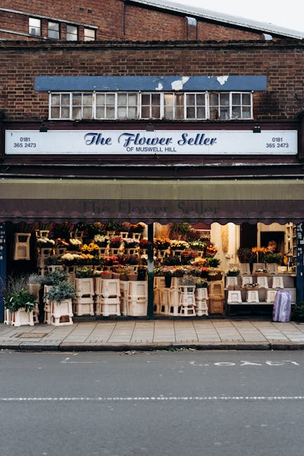 The exterior view of The Flower Seller shop located on Muswell Hill, featuring a traditional brick facade and a prominent blue awning with the store's name in elegant script. Below the sign, a large d