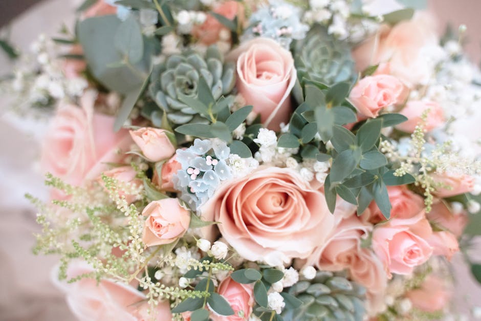 A floral arrangement featuring large, soft pink roses with tightly packed petals, complemented by smaller pale pink rosebuds. Intertwined with pale green succulent leaves and sprigs of delicate white 