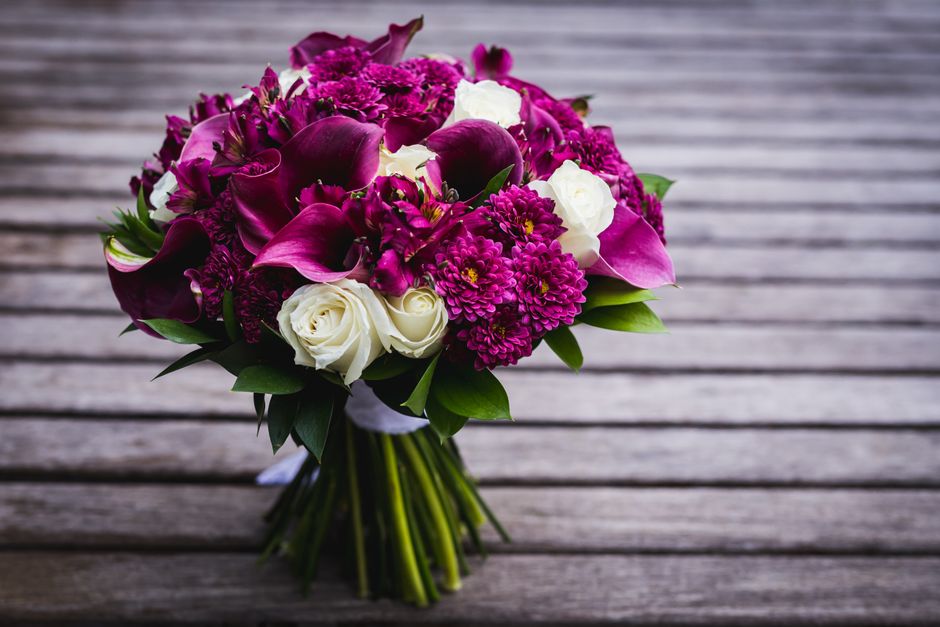 A fresh floral bouquet featuring deep purple calla lilies, magenta dahlias, and white roses, arranged in a compact, rounded style with lush green leaves interspersed among the flowers. The bouquet is 
