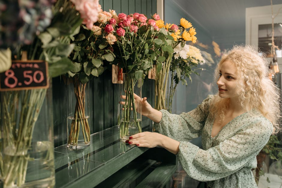 A young woman with curly blonde hair and fair skin selecting a floral gift at a florist shop. She is wearing a light, patterned blouse with long, puffed sleeves and has red painted nails. She is stand