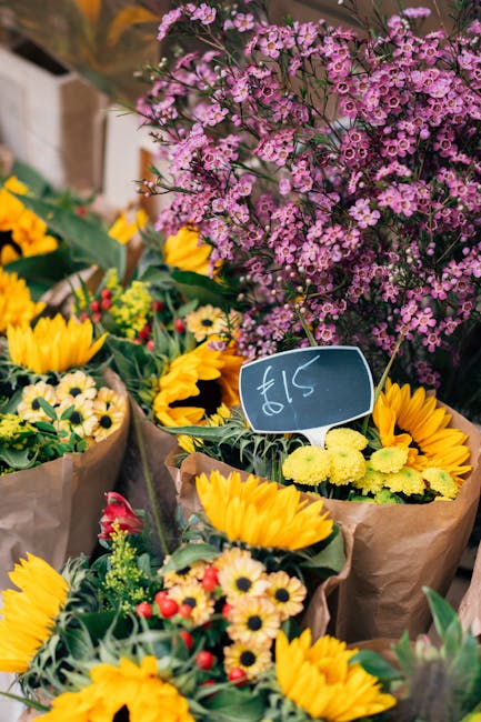A vibrant display of freshly arranged floral bouquets featuring bright yellow sunflowers, small yellow daisies, and red and white flowers wrapped in brown paper, positioned outdoors on a table. In the