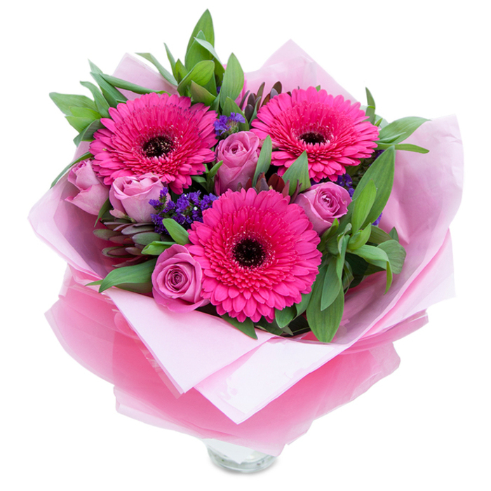 I arranged this bouquet to balance boldness and tenderness, photographed here against a pure white backdrop so the colours feel immediate and honest. Three vivid fuchsia Gerbera daisies dominate the scene, their layers of slender petals radiating outward and each centred by a dark, nearly black eye that grounds the composition. Nearby, several roses in a muted lavender-pink or dusty-rose are tightly budded, their soft, velvety curves suggesting intimacy and freshness. Pockets of deep purple statice are tucked between the larger blooms, creating tiny islands of texture and a cool tonal foil to the warm pinks. The greenery - broad salal-like leaves and the grey-green hint of eucalyptus - provides a structured yet dewy frame, adding a fresh, almost herbal lift to the scent. The bouquet is wrapped with care: an inner sheet of soft pale pink tissue and an outer layer of translucent fuchsia-pink cellophane, slightly crinkled so it catches the light and feels celebratory without being fussy. At the base, the clear glass vase rim peeks through, indicating the arrangement is prepared for immediate display in a home or office. The effect is romantic without being overwrought, playful while still refined - a suitable choice for birthdays, anniversaries, or a warm 'thank you' left on a doorstep. As a local florist familiar with Golders Green and the quieter streets off Golders Green Road, I imagine this bouquet arriving to lift someone's day, its colours bright against a London interior and its scent offering a small, comforting pause.